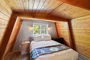 A bedroom with a sloped ceiling, a bed with white bedding, a bedside table, a nightstand, and a window with a view of greenery outside. - Ski-lee Retreat Ski Hike Explore Sleeps 10 (Brian Head)