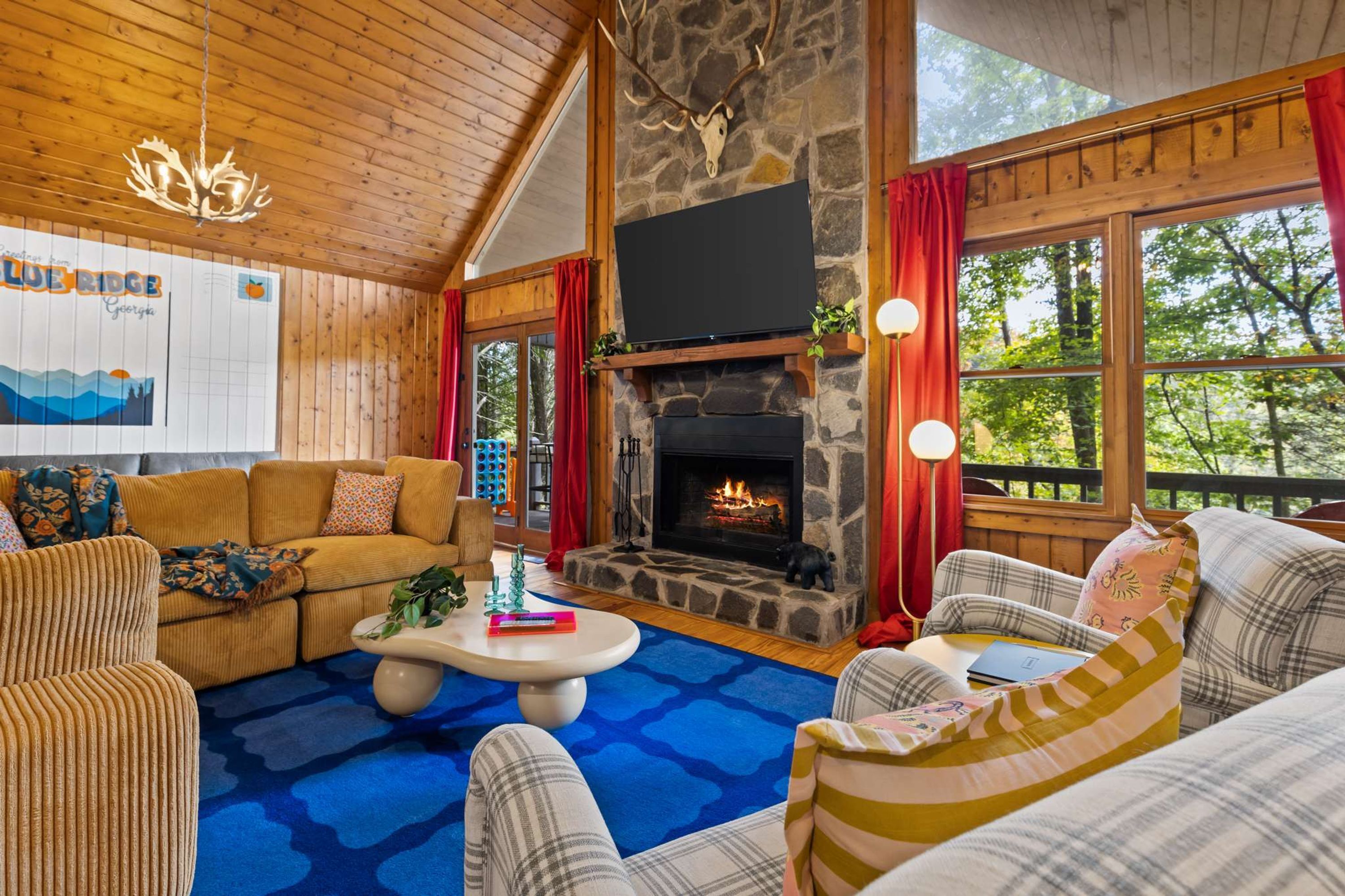 A cozy living room with a stone fireplace, wooden ceiling, and large windows.