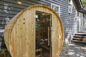 Estructura de sauna de madera con puerta de vidrio, un escalón de piedra que la precede y un banco de madera en su interior.
