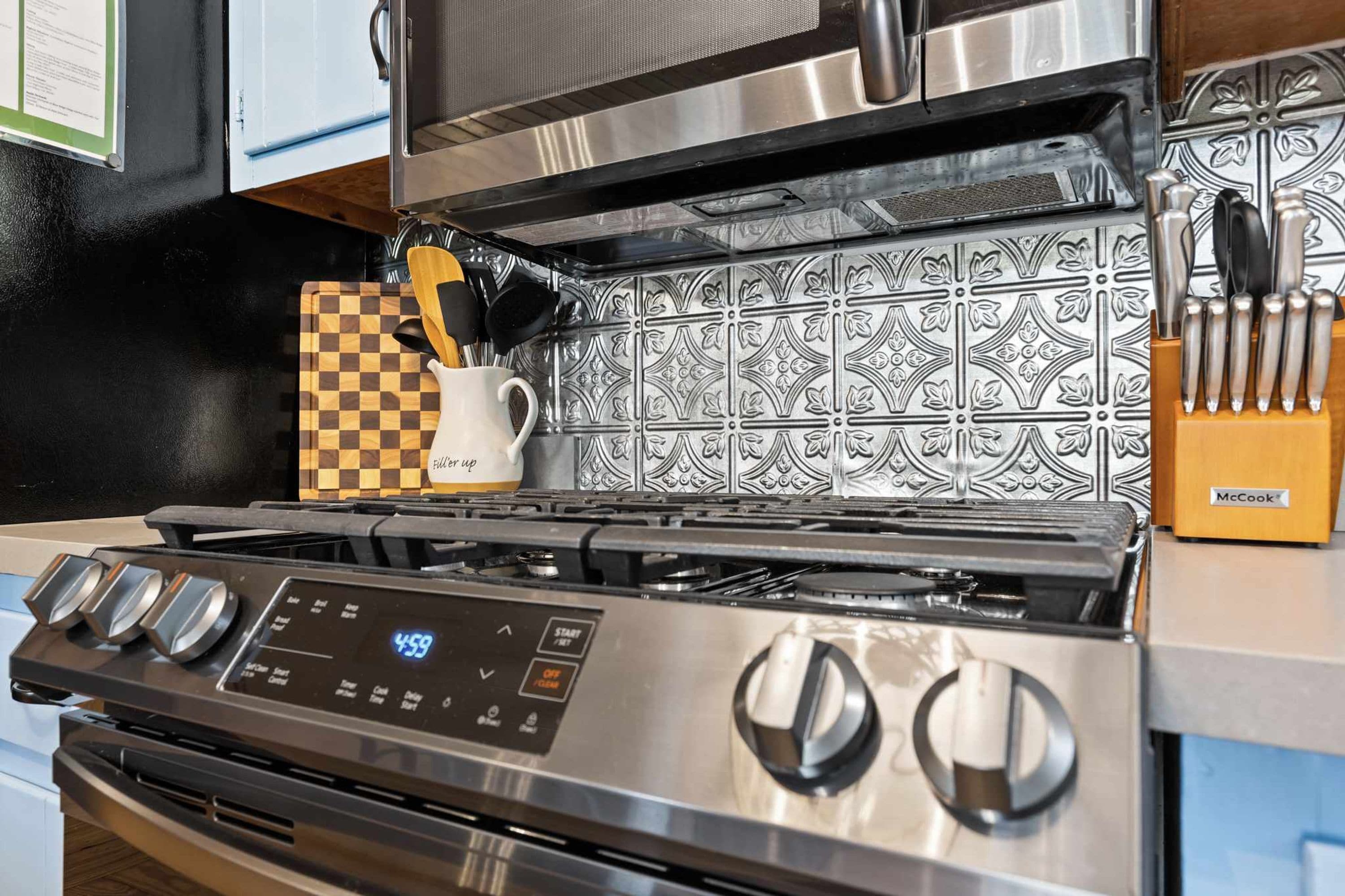 A stainless steel range with a digital control panel, a checkered cutting board, a white mug, and a knife block.