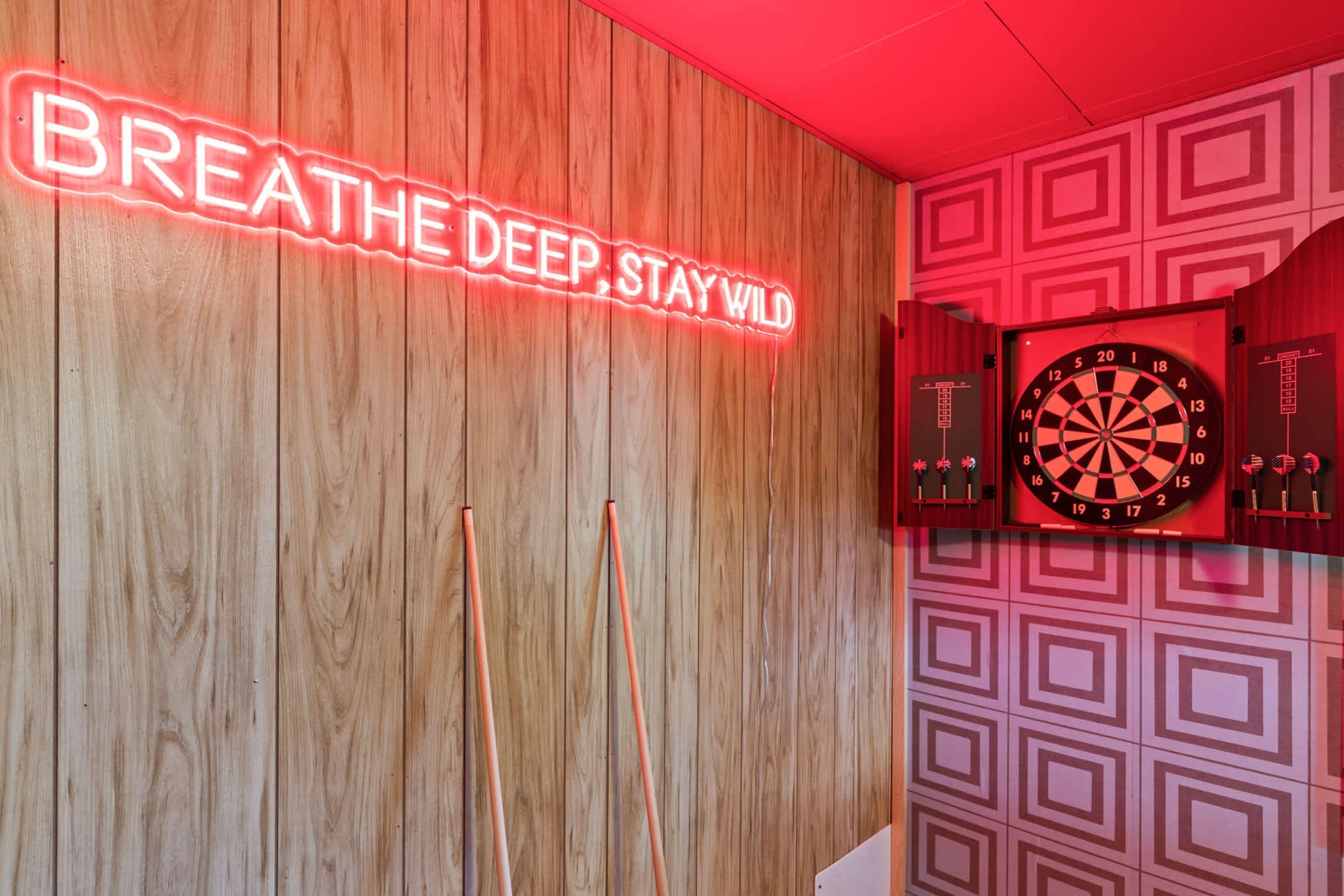 Een dartbord aan een muur, met een neonbord erboven met de tekst 'BREATHE DEEP, STAY WILD'.