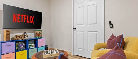 A living room with a television displaying 'NETFLIX,' a bookshelf with children's books, a chessboard, and a yellow armchair.