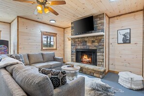 Un salon chaleureux avec une cheminée en pierre, un téléviseur à écran plat fixé au-dessus et un ventilateur de plafond.