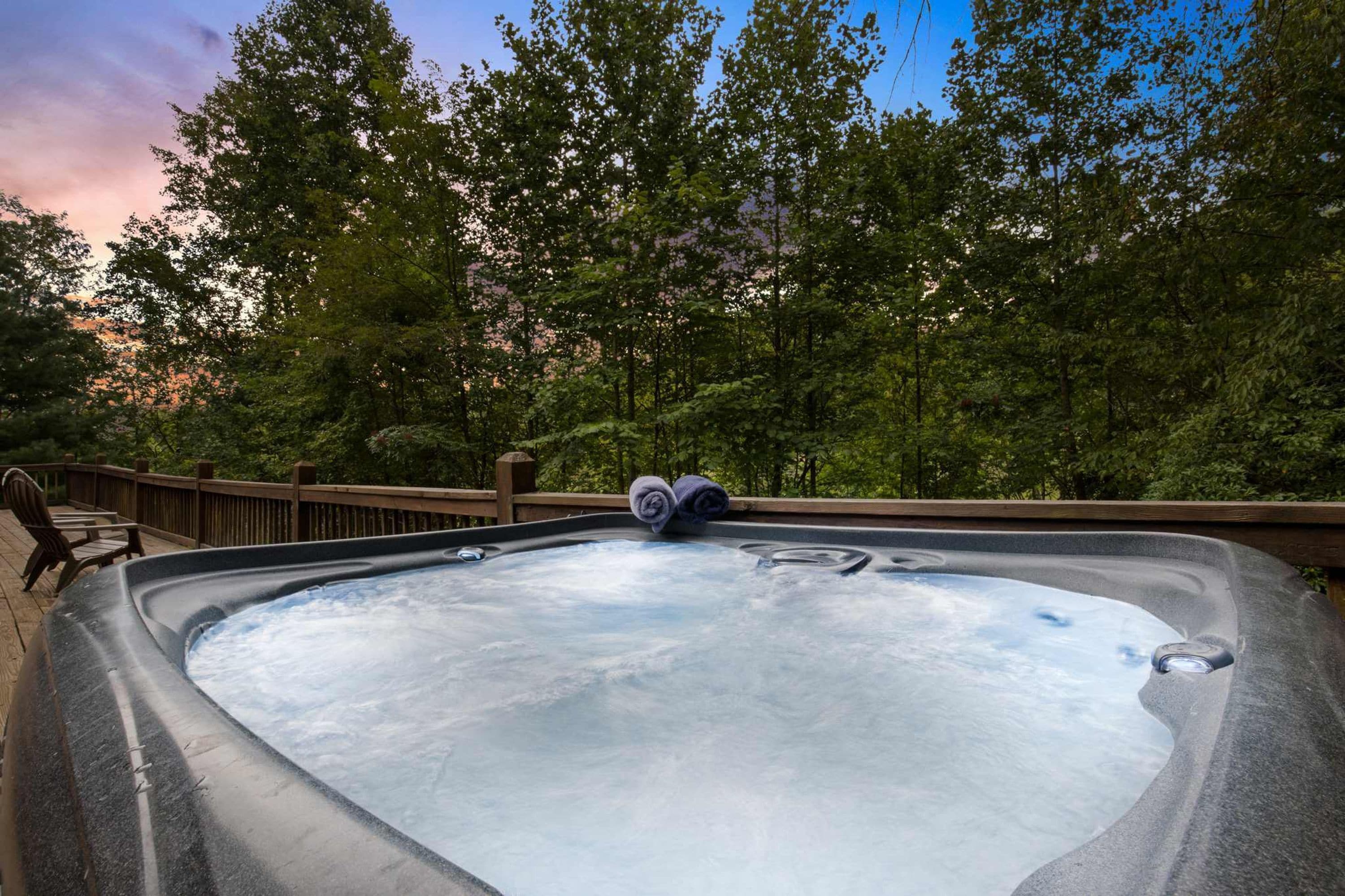 A hot tub surrounded by a wooden deck with chairs, set against a backdrop of trees and a sunset sky.