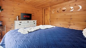 A wooden room with a bed, a white dresser, a television displaying the Netflix logo, and a wall with crescent moon cutouts. - Cozy Mountain Escape Hot Tub Fire Pit Views (Blue Ridge)