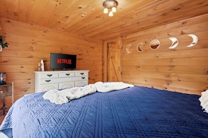 Ein Zimmer mit Holzboden, einem Bett, einer weißen Kommode, einem Fernseher, der das Netflix-Logo zeigt, und einer Wand mit Mondsichel-Aussparungen.