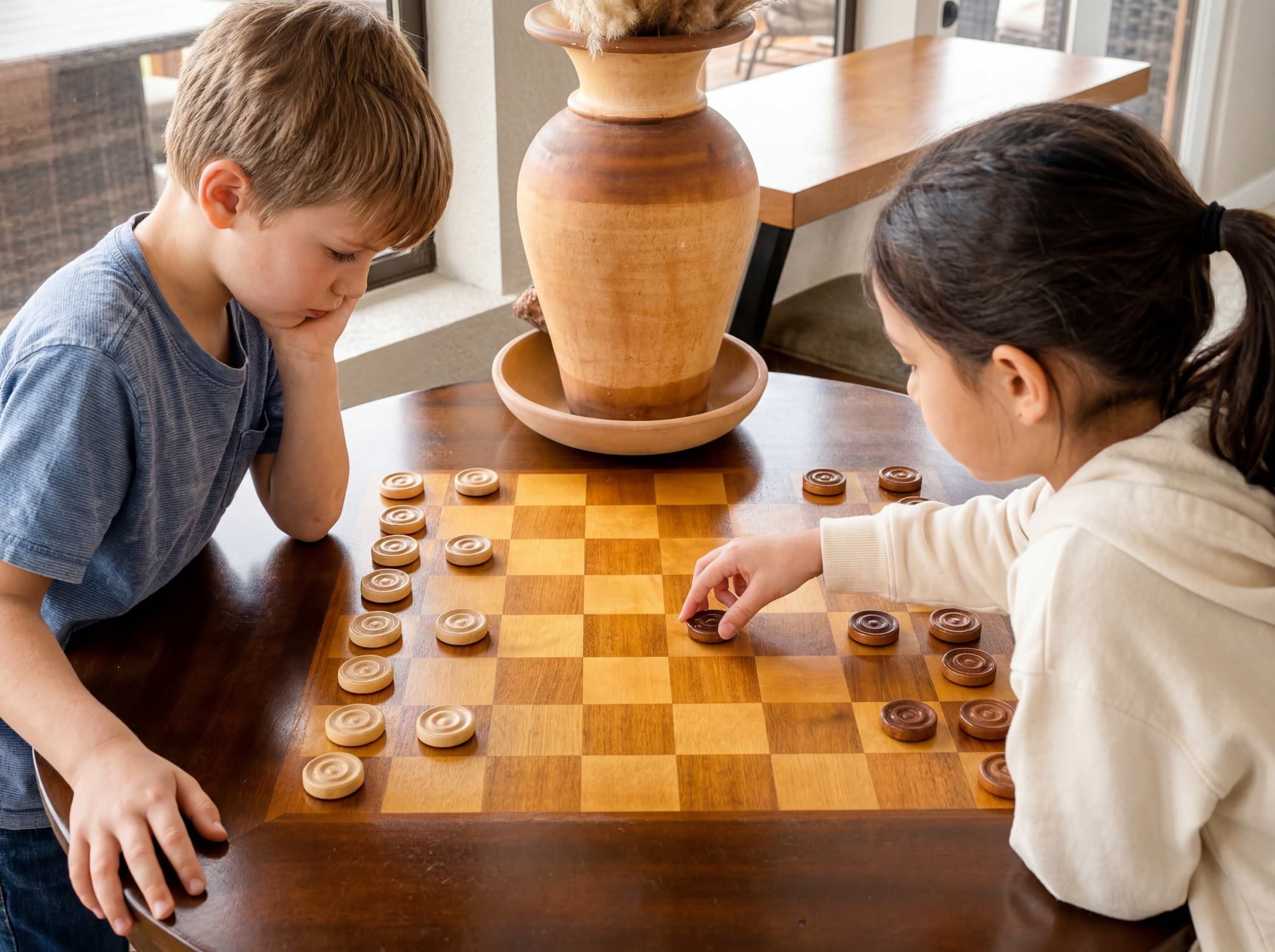 Two children playing chess on a wooden table with a large vase in the background.
