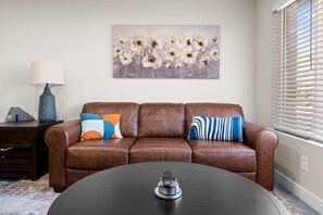 Ein Wohnzimmer mit braunem Ledersofa, einem runden Couchtisch, einer Lampe, einem Gemälde und einem Fenster mit Jalousien.