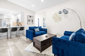 A modern living room with blue sofas, a wooden coffee table, and a kitchen in the background. - Poolside Patio Close to Westgate Dog Friendly (Phoenix)