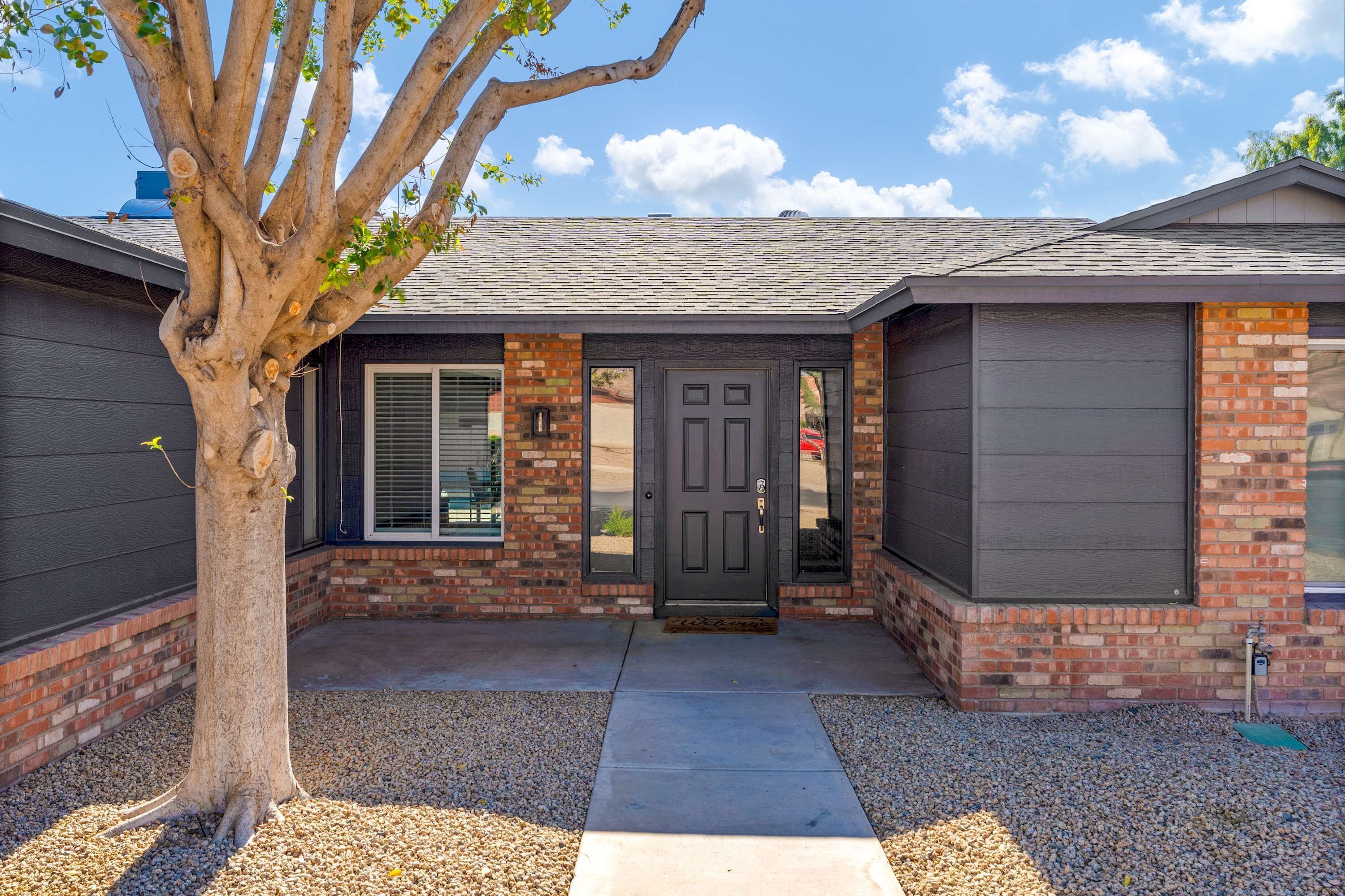 A residential house with a brick facade, a dark front door, and a tree in the front yard.