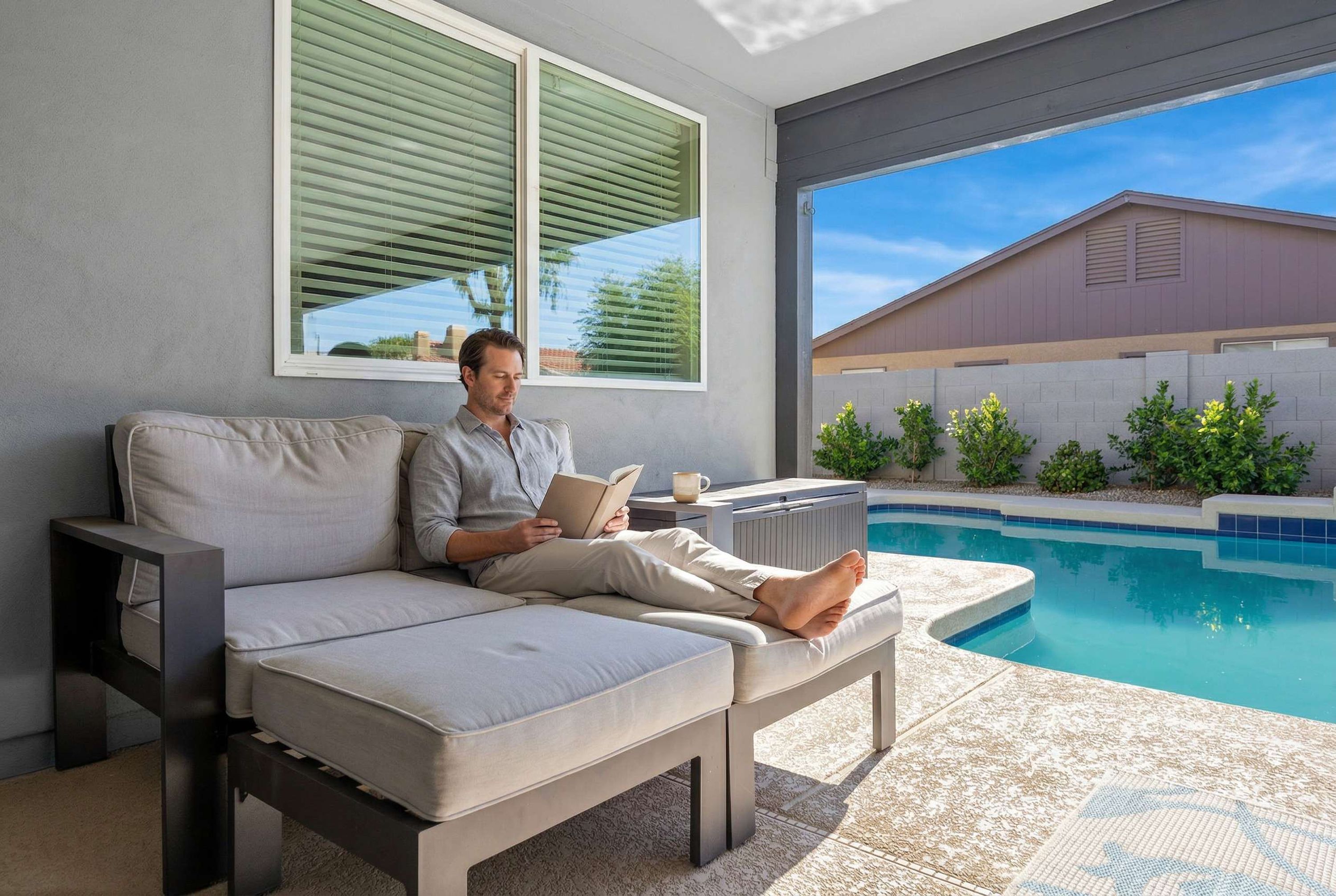 Ein Mann entspannt auf einer Couch am Pool mit Blick auf ein Haus und Grünflächen.