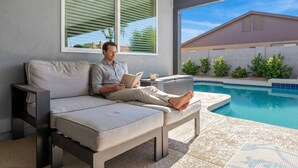 Ein Mann entspannt auf einer Couch am Pool mit Blick auf ein Haus und Grünflächen.
