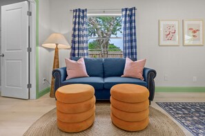 A living room with a blue sofa, two round ottomans, a lamp, and a view of a tree through a window with checkered curtains. - Resort Style Backyard Tiki Bar Renovated Pool (Largo)