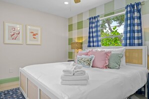 A neatly made bed with white linens and a stack of folded towels, a window with blue and white checkered curtains, and two framed wall art pieces. - Resort Style Backyard Tiki Bar Renovated Pool (Largo)