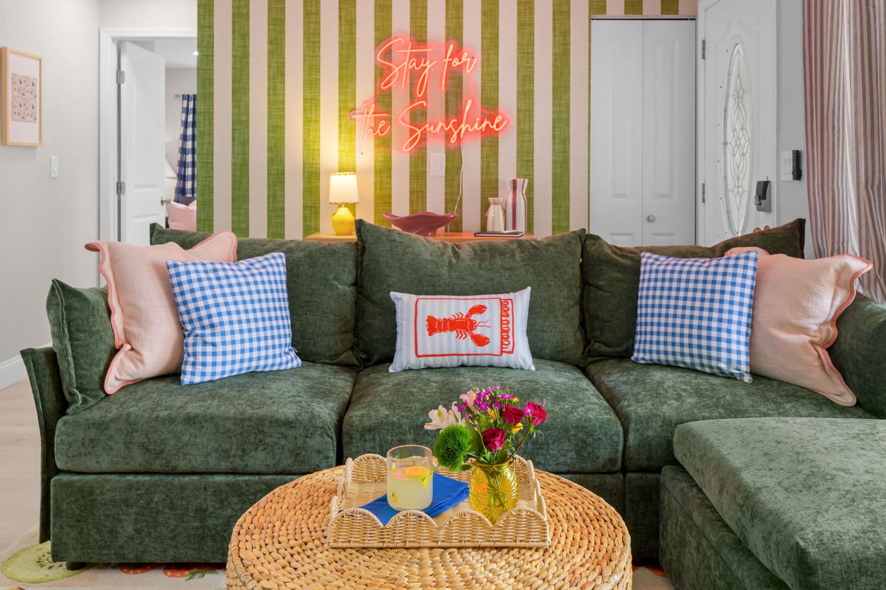 Un sofá verde con almohadones a cuadros, una mesa de centro trenzada con una bebida y flores, y un letrero de neón que dice 'Stay for the Sunshine' en una habitación con papel tapiz a rayas.