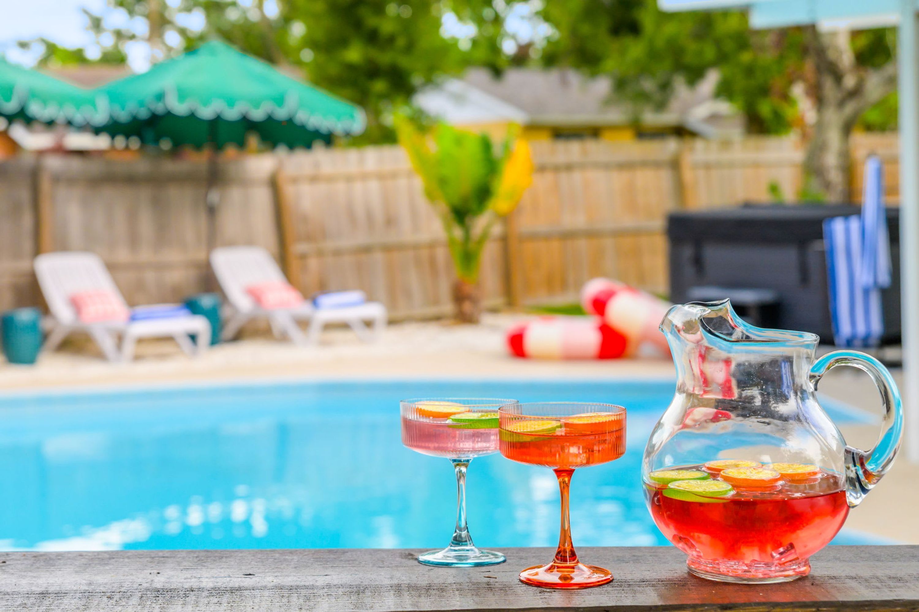 A poolside setting with a clear view of the pool, lounge chairs, and a pitcher of a beverage with fruit slices.