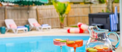 A poolside setting with a clear view of the pool, lounge chairs, and a pitcher of a beverage with fruit slices.