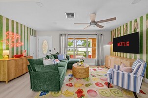 A living room with a green sofa, a striped armchair, a flat-screen TV, and a neon sign.