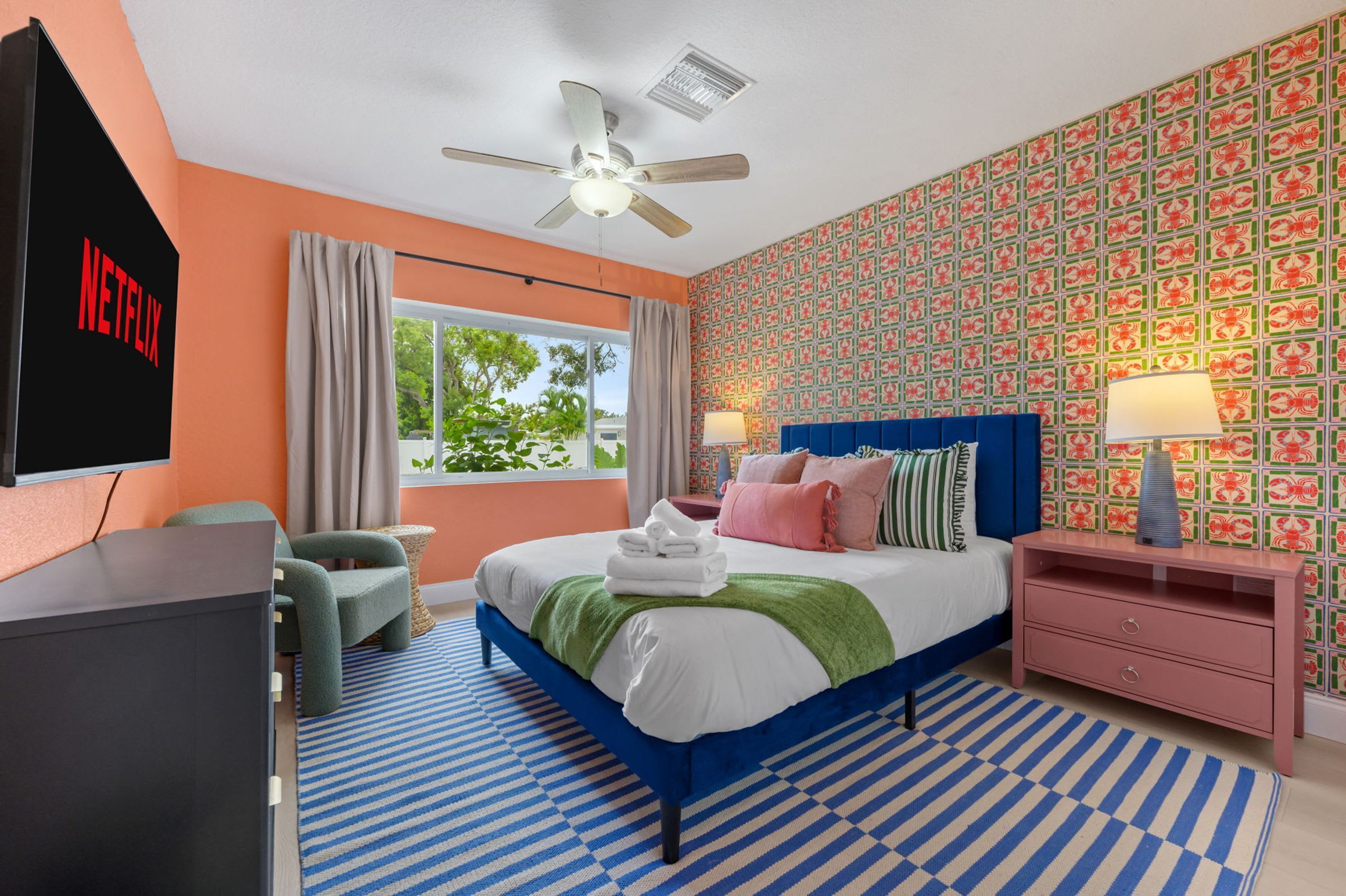 Una habitación de hotel moderna con una cama, un televisor, un ventilador de techo, una ventana con cortinas, un papel tapiz estampado y una alfombra a rayas.