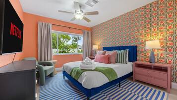 Una habitación de hotel moderna con una cama, un televisor, un ventilador de techo, una ventana con cortinas, un papel tapiz estampado y una alfombra a rayas.