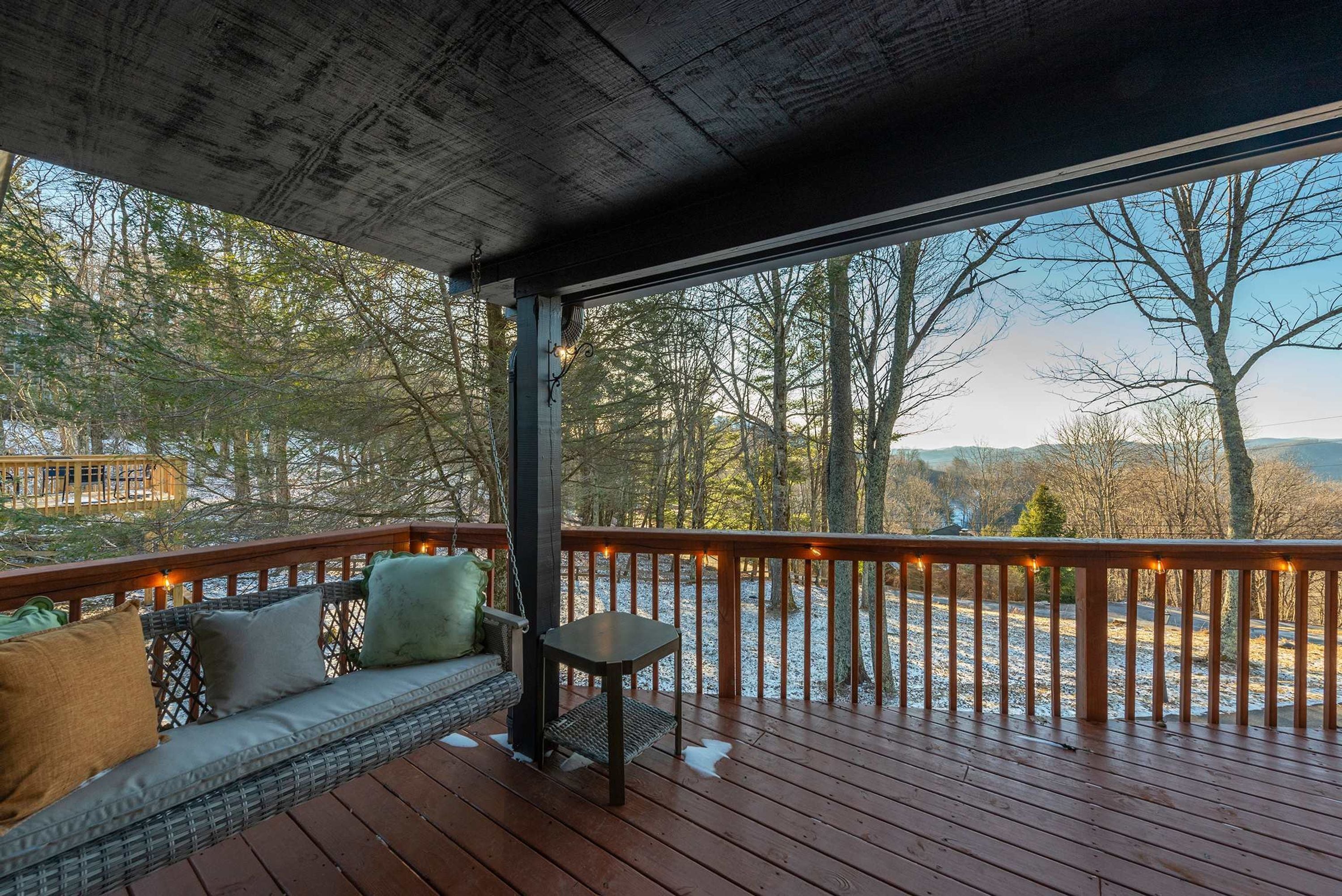 A covered outdoor deck with a wooden railing, a bench with cushions, and a view of a snowy landscape and trees.