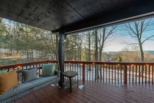 A covered outdoor deck with a wooden railing, a bench with cushions, and a view of a snowy landscape and trees.