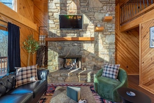 A cozy interior with a stone fireplace, leather sofas, a green armchair, and a flat-screen TV mounted above the fireplace.