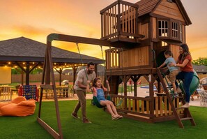A wooden play structure with a swing set and climbing elements, surrounded by a grassy area and a covered seating area. - 6k Sqft Estate - Epic Amenities Fun - Sleeps 22 (San Antonio)