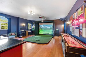 A room with a golf simulator setup, including a screen displaying a golf course, a putting green, and a wooden table with golf balls. - 6k Sqft Estate - Epic Amenities Fun - Sleeps 22 (San Antonio)