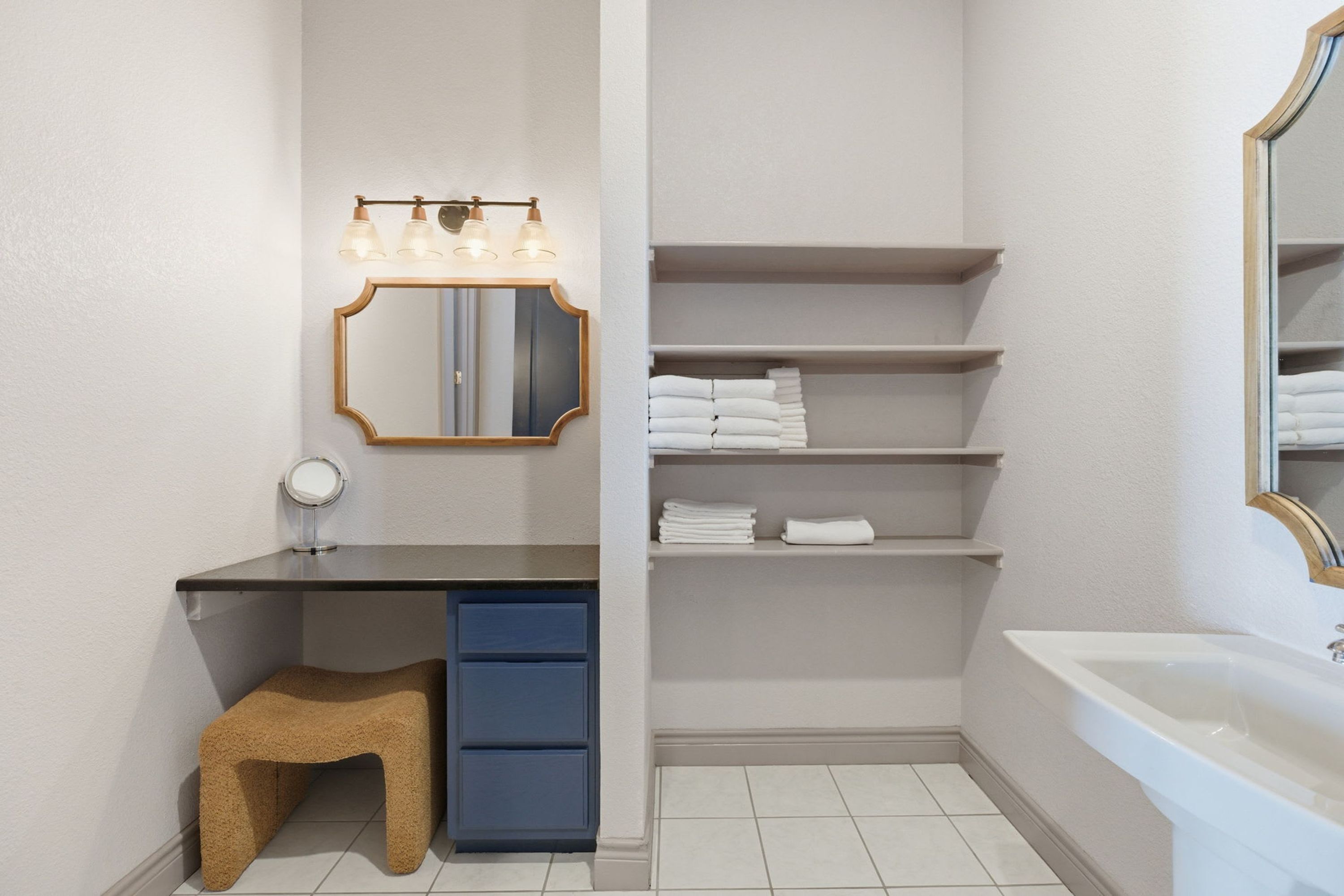 Un baño con tocador, un taburete, estantes con toallas y una bañera.