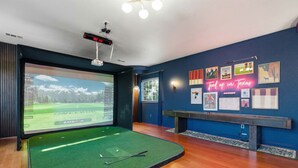A room with a large screen displaying a golf course, a pool table, and a wall with various framed pictures and a neon sign. - 6k Sqft Estate - Epic Amenities Fun - Sleeps 22 (San Antonio)