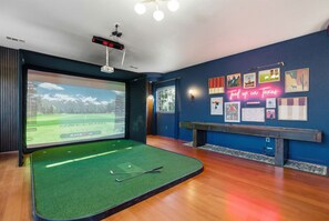 Ein Zimmer mit einem großen Bildschirm, der einen Golfplatz, ein Billardtisch und eine Wand mit verschiedenen gerahmten Bildern sowie eine Neonreklame zeigt.