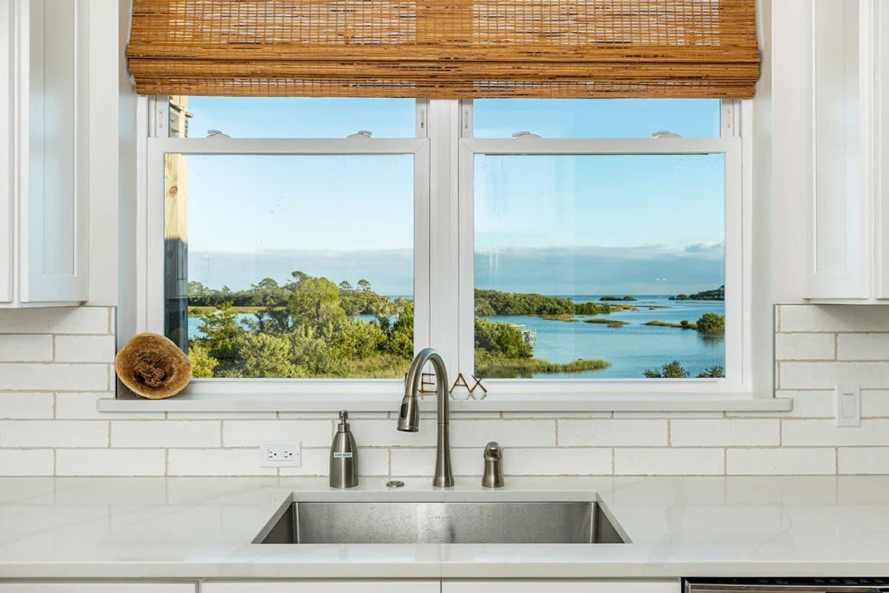 A kitchen with a double sink, a window with a view of a natural landscape, and a bamboo blind.