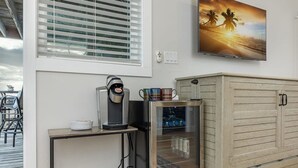 A room with a coffee maker on a small table, a cabinet with a glass door containing beverages, and a painting of a sunset on the wall. - 3 Story Haven Fire Pit Kayaks Updated 360 Views (Cedar Key)