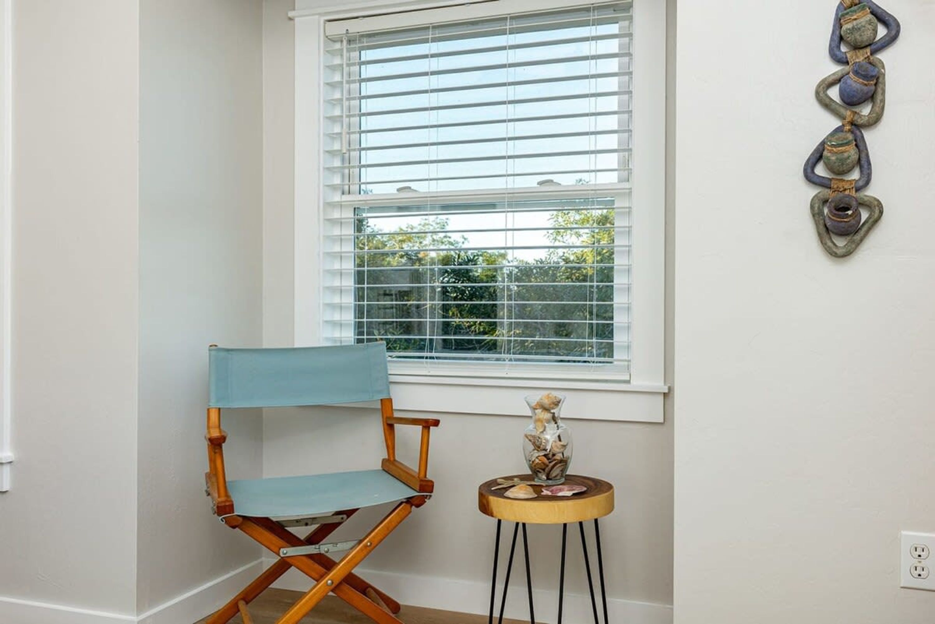 Una habitación con ventana, una silla plegable azul y una pequeña mesa de madera con un jarrón de conchas marinas.