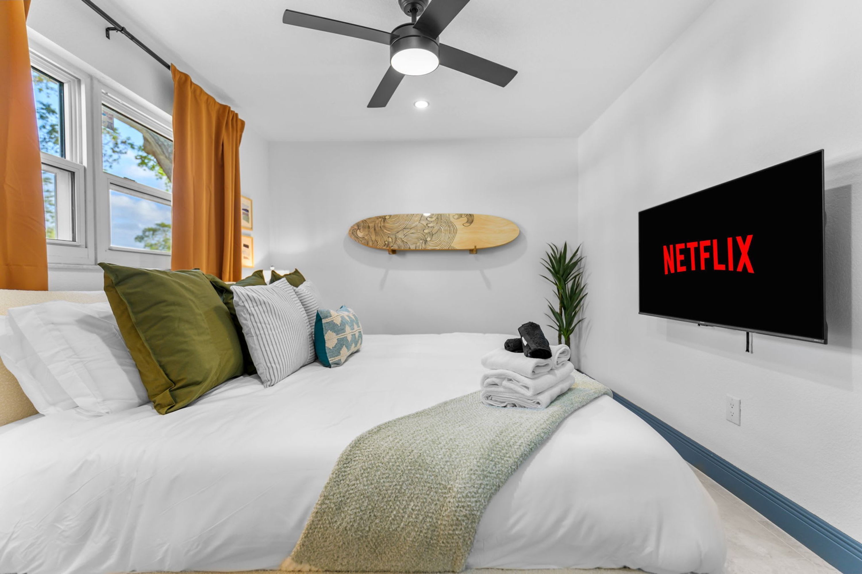 A hotel room with a bed, a ceiling fan, a mounted TV, and a wall-mounted shelf with a skateboard.