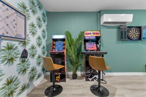 Quarto com duas estações de jogos de fliperama, um alvo de dardos e um jogo de palavras na parede.