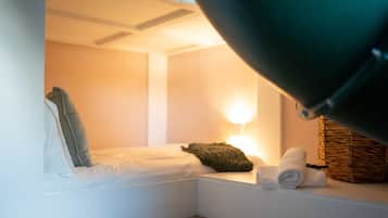 Um quarto pequeno e aconchegante com uma cama, um travesseiro, uma luminária e um cesto.
