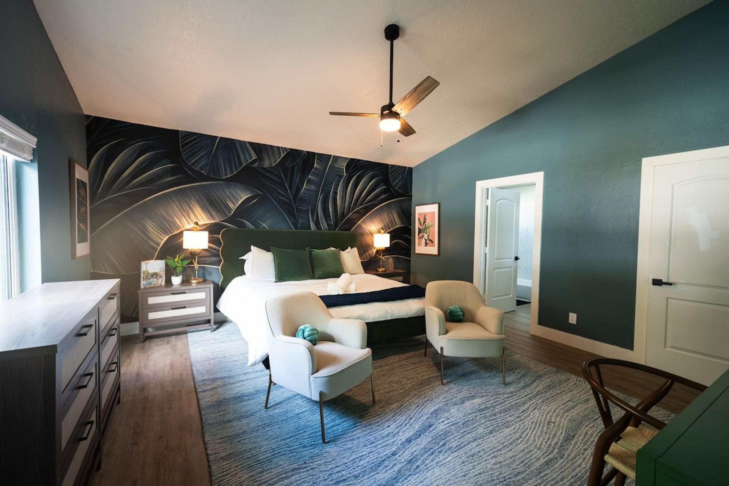 A bedroom with a bed, two chairs, a nightstand, a ceiling fan, and a door.