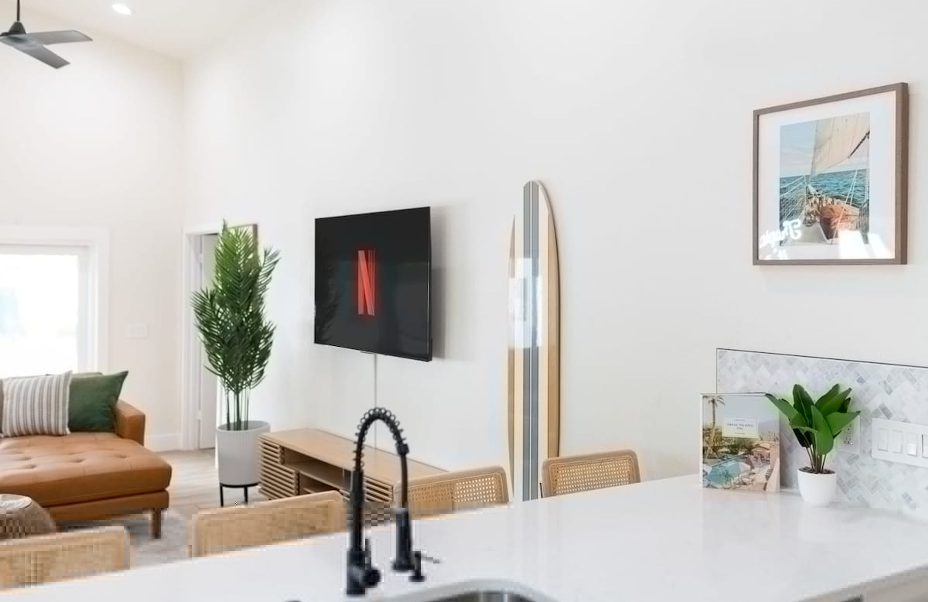 Un espacio habitable moderno con un televisor de pantalla plana montado en la pared, una tabla de surf apoyada contra la pared y una zona de comedor con una mesa y sillas blancas.