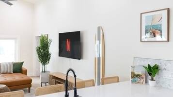 Un espacio habitable moderno con un televisor de pantalla plana montado en la pared, una tabla de surf apoyada contra la pared y una zona de comedor con una mesa y sillas blancas.