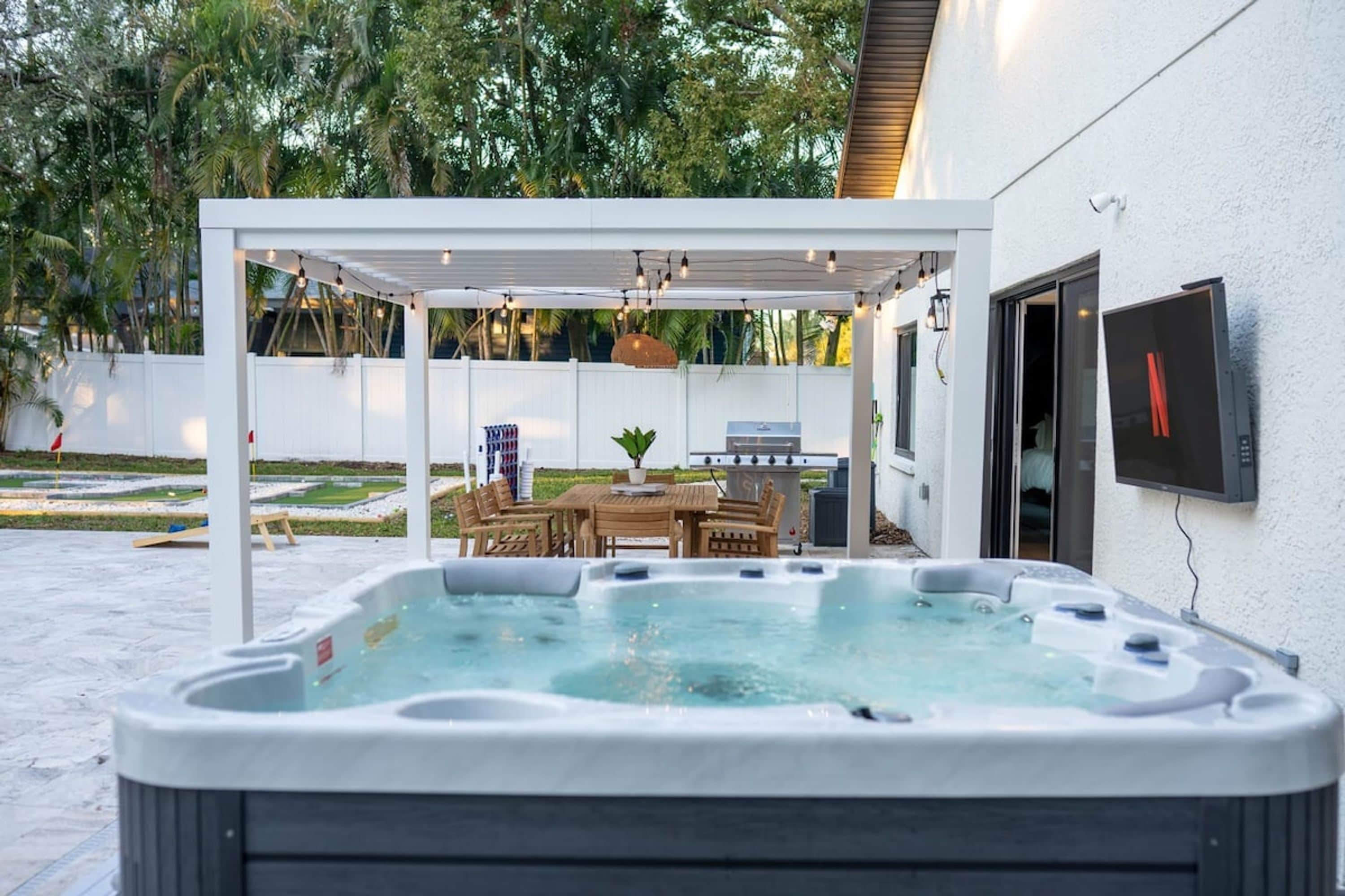 A hot tub with a pergola overhead, a barbecue grill, and a dining area with outdoor furniture.