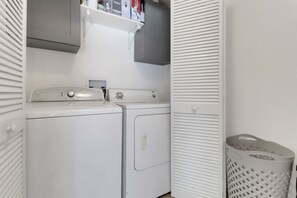 A laundry area with a washing machine and dryer, a shelf above the dryer, and a laundry basket. - High Tide - 3 Br Sleeps 6 - Private Pool - Games (Naples)