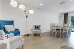 Ein modernes Wohnzimmer mit einem blauen Sofa, einem weißen Sessel, einem Esstisch mit Stühlen und einer Stehlampe.