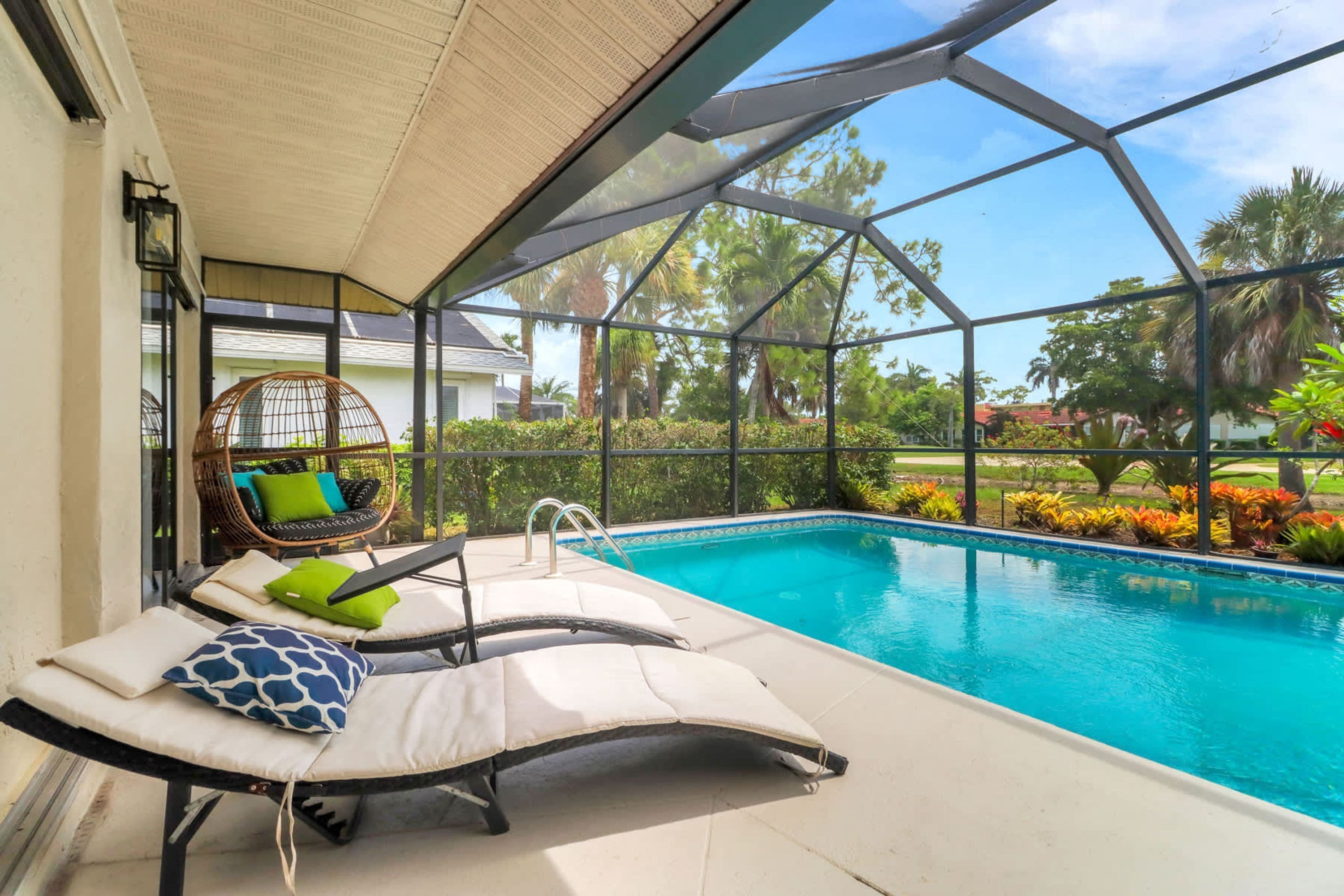 A poolside area with lounge chairs, a wicker chair, and a view of the pool and surrounding greenery.