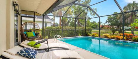 A poolside area with lounge chairs, a wicker chair, and a view of the pool and surrounding greenery.