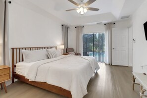 Ein Schlafzimmer mit einem Holzbett, einem Nachttisch, einem Schreibtisch, einem Deckenventilator und Blick auf einen Pool durch eine Glastür.