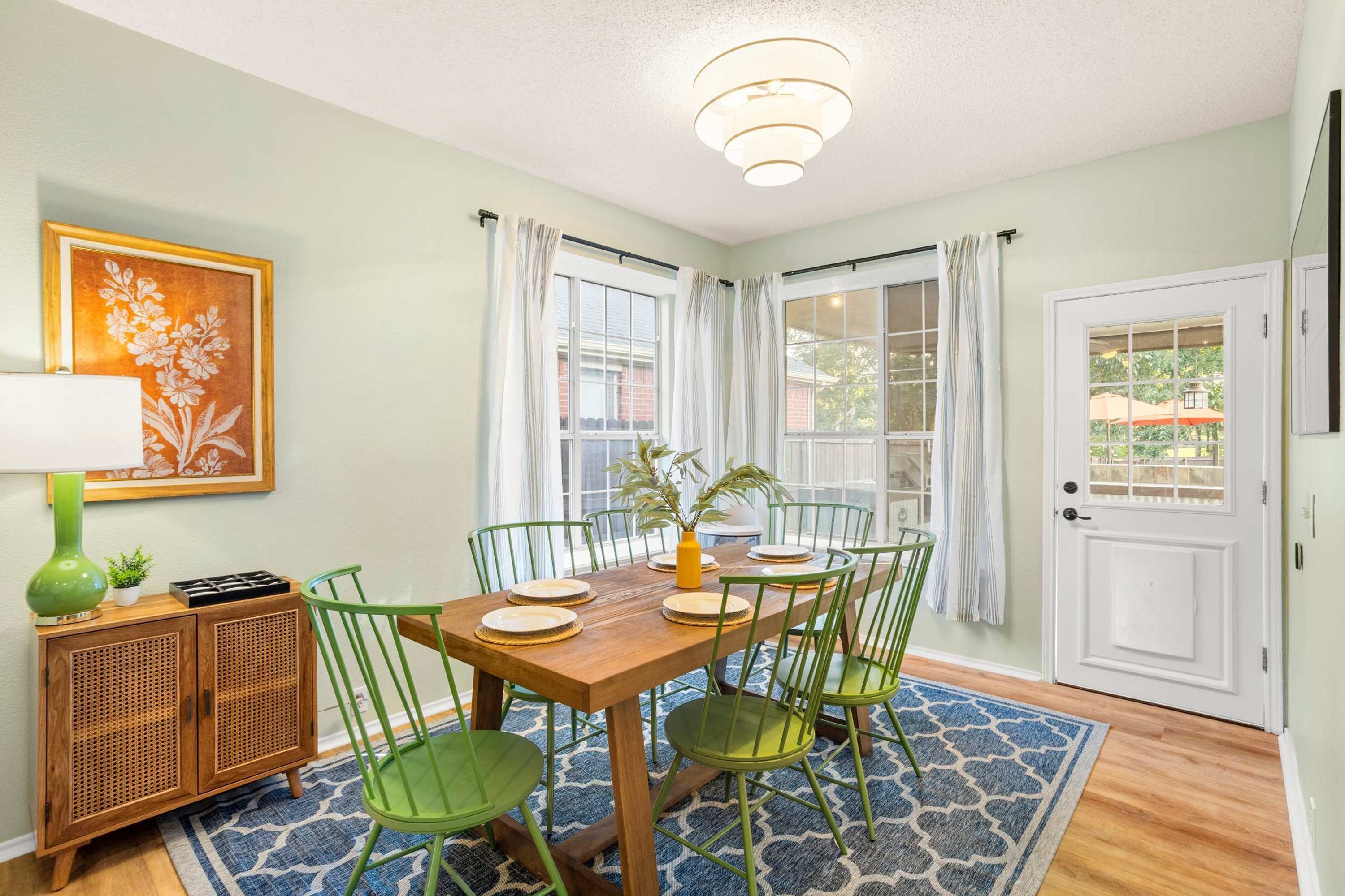 A dining room with a wooden table set for four, green chairs, a rattan cabinet, a green vase, and a framed floral painting.