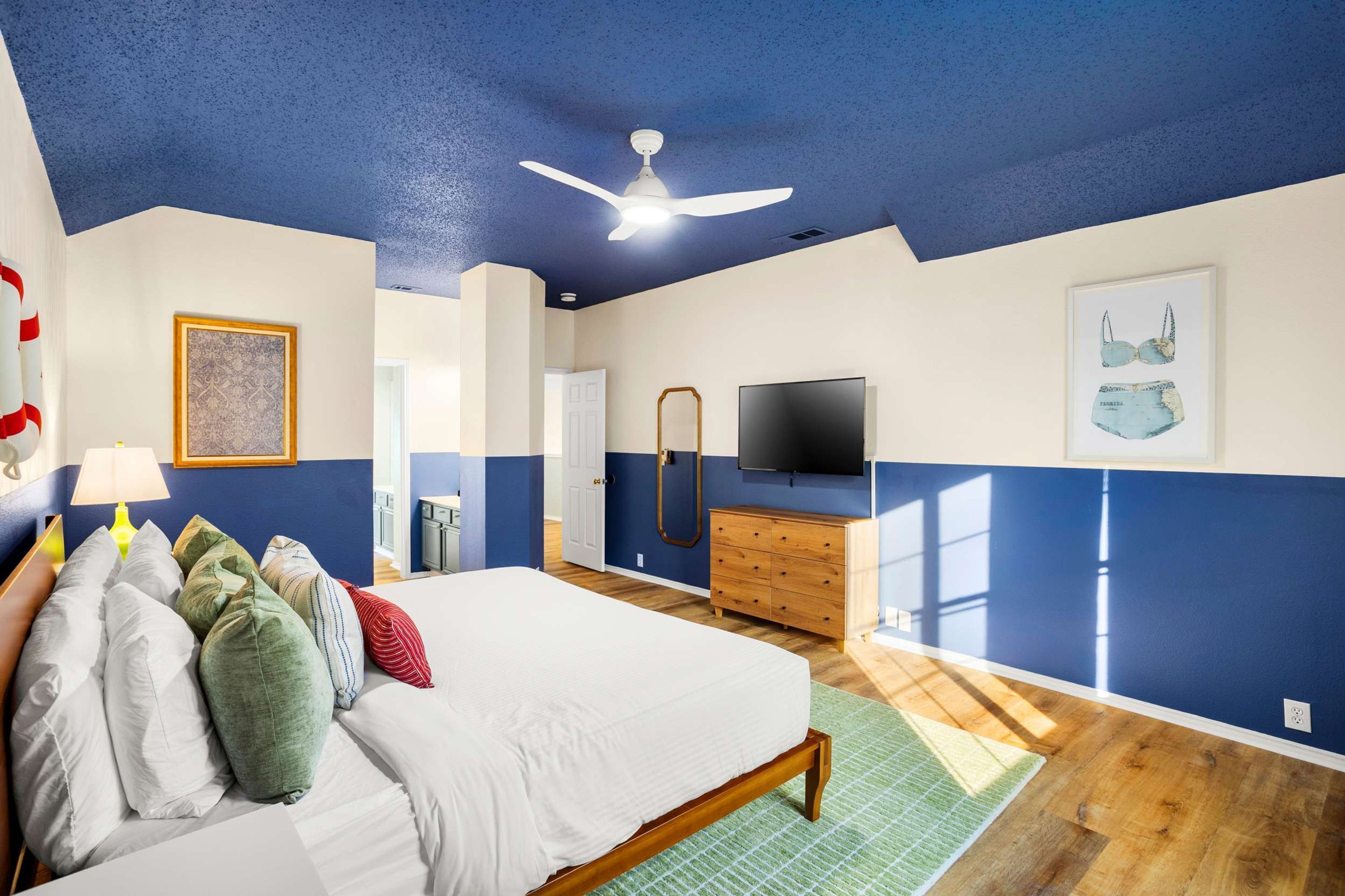 Een slaapkamer met een bed, kussens, een dressoir, een televisie en een plafondventilator.