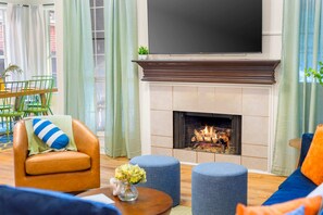 Ein Wohnzimmer mit Kamin, einem flachbildfernseher darüber, einem Ledersessel, Poufs und einem Essbereich mit Stühlen und einem Tisch.